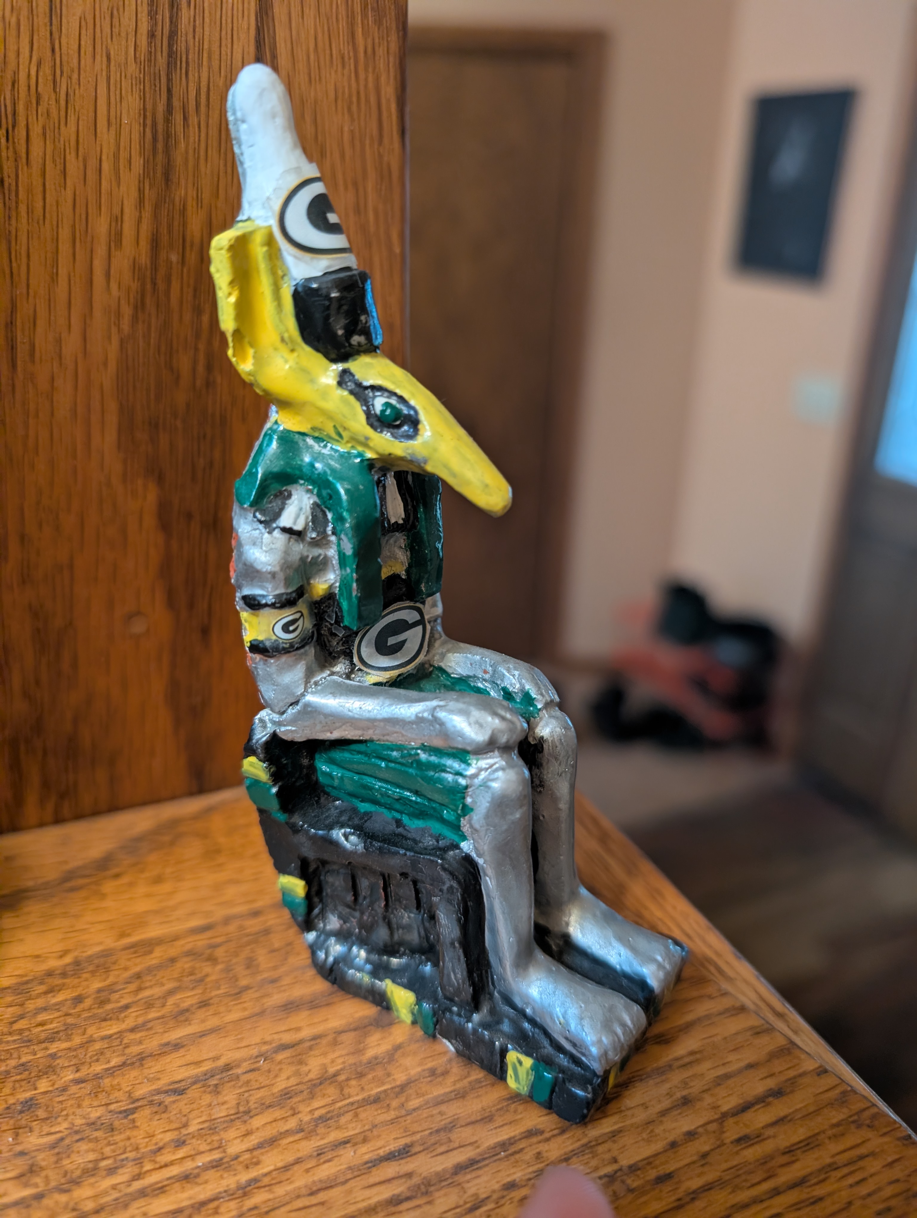 A plaster statuette of Set wearing a double crown. He is painted with a yellow face and green hair, with other green and gold details, and has Green Bay Packer decals stuck to him.